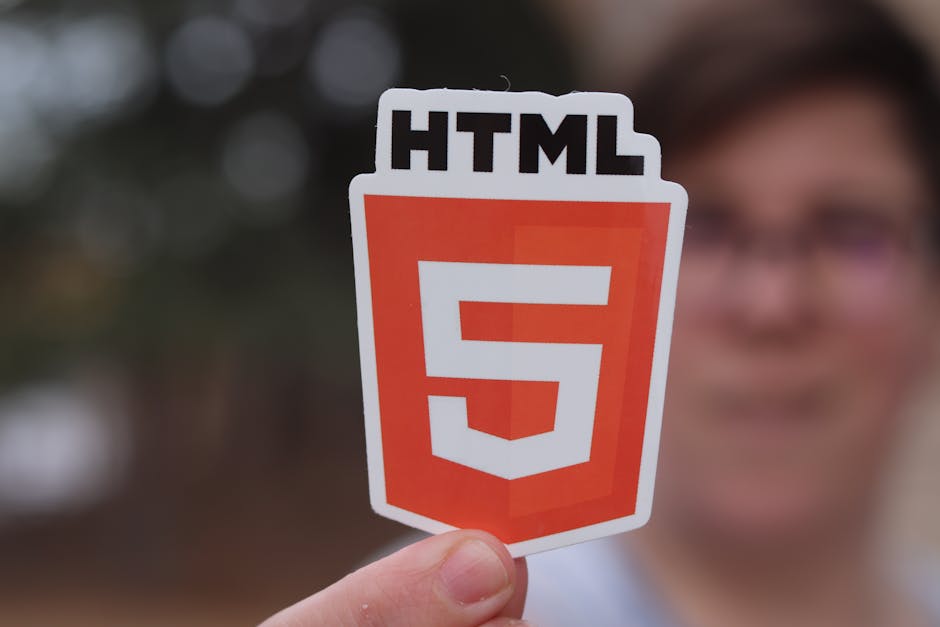 A person holding a sticker with the HTML5 logo, featuring a white shield shape with a bold red background and the number five in white at its center. The person's hand is visible, with short nails, an